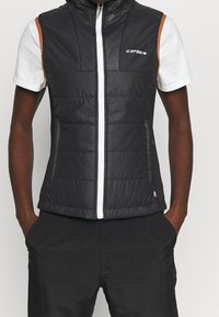 Svart pufferväst med hög krage, vit dragkedja och orange detaljer; har quiltad textur och sidofickor.