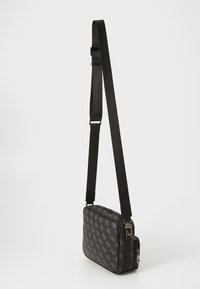 Black crossbody bag with adjustable strap, patterned with a repeated light gray monogram, and two zippered compartments on a white background.