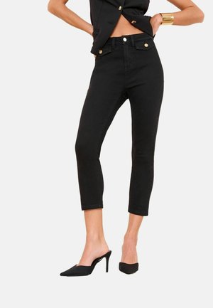 Femeie purtând blugi negri tăiați, cu detalii de nasturi aurii, asortați cu pantofi mule negri cu vârf ascuțit și un top negru fără mâneci.