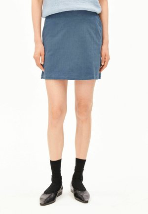Frau trägt einen blauen Cord-Minirock, hellblaues Oberteil, schwarze Socken und schwarze flache Schuhe und steht vor weißem Hintergrund.