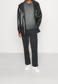 Chaqueta negra, brillante y a prueba de agua con capucha, combinada con una camisa de manga larga gris con textura y pantalones oscuros. Zapatillas blancas completan el atuendo.