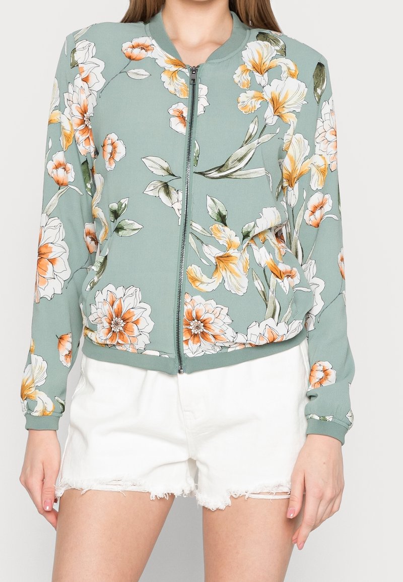 Blouson aviateur floral vert clair, avec des fleurs orange et blanches, des poignets côtelés et une fermeture éclair à l'avant. Associé à un short blanc effiloché.