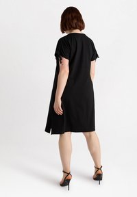 Czarna sukienka z krótkim rękawem o luźnym kroju, z bocznymi rozcięciami i zawiązywanymi rękawami. Noszona z czarnymi sandałami na wysokim obcasie. Gładka tekstura materiału.