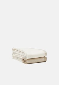 Deux serviettes en coton pliées empilées, une crème dessus et une beige en dessous, toutes deux avec des bords frangés.