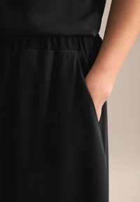 Schwarzes Kleidungsstück mit einer glatten Textur, das einen elastischen Bund und seitliche Taschen aufweist. Der Stoff wirkt leicht und weich.