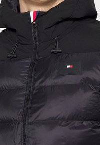 Czarna kurtka puchowa z wysokim kołnierzem, z zamkiem błyskawicznym i troczkami. Posiada małą naszywkę z logo w kolorach czerwonym, białym i niebieskim.