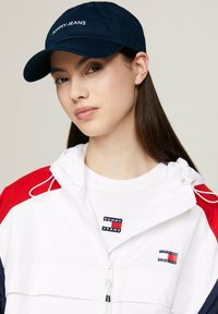 Granatowa czapka z haftem "TOMMY JEANS" w kolorze białym. Noszona z białą kurtką z czerwonymi i granatowymi akcentami, na tle białego T-shirta z logo.