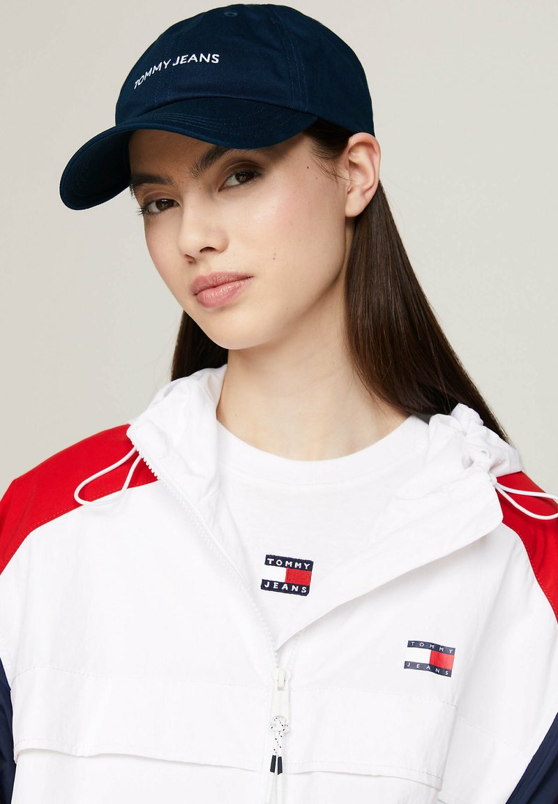 Granatowa czapka z haftem "TOMMY JEANS" w kolorze białym. Noszona z białą kurtką z czerwonymi i granatowymi akcentami, na tle białego T-shirta z logo.