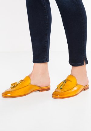 Osoba w błyszczących żółtych skórzanych mokasynach bez pleców ozdobionych frędzlami, w parze z ciemnoniebieskimi dopasowanymi dżinsami na białym tle.