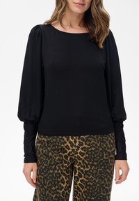 Kobieta ubrana w czarną bluzkę z długim rękawem i bufkami oraz spodnie w panterkę, stojąca na tle białym.