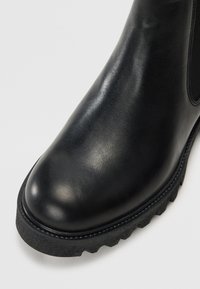 Czarne skórzane botki za kostkę z błyszczącym wykończeniem, zaokrąglonym noskiem i teksturowaną gumową podeszwą z wzorem w zygzak. Widać boczne elastyczne panele.