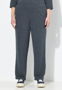 Person med lösa, mörkgrå, strukturerade byxor och marinblå sneakers med vita sulor och snören, som står mot en vit vägg.