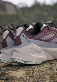 Ein Paar mauvefarbene und graue Adidas Terrex Trailrunning-Schuhe, die auf felsigem Gelände im Freien ruhen.