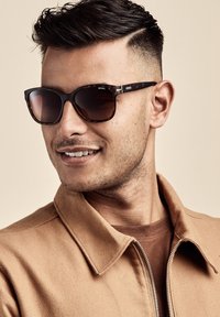 Ruskeat aurinkolasit tummilla linsseillä, suorakulmainen muoto ja kilpikonnakuosi. Yhdistettynä vaaleanruskeaan takkiin ja ruskeaan paitaan.