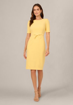 Kvinde iført en tætsiddende gul knælængde kjole med korte ærmer, et bælte i taljen og beige højhælede sandaler, stående mod en ensfarvet baggrund.