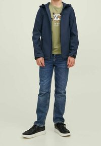 Child wearing a navy jacket, green t-shirt with a dog graphic, blue jeans, and black sneakers with white soles, standing with hands relaxed.