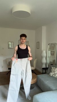 Svetlošedé cargo nohavice s viacerými bočnými vreckami, vo voľnom strihu, s pripevnenou etiketou, ktoré drží osoba v čiernom tielku.