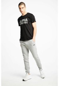 T-shirt en coton noir avec "ALPHA INDUSTRIES" imprimé en blanc, associé à un pantalon de jogging gris et des chaussures de sport blanches avec des accents noirs.