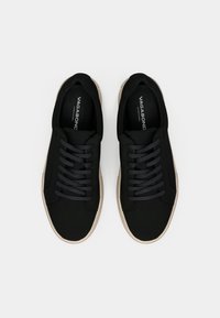 Încălțăminte sport neagră cu o textură netedă, vârf rotund, șireți plat și o talpă de contrast bej. Logo-ul "VAGABOND" pe branț.