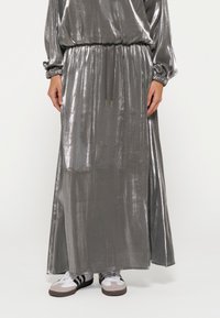 Maxi dress grigio metallizzato con maniche lunghe, vita arricciata e dettaglio con cordoncino. Accostato a calzature bianche e accenti grigi.