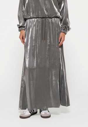 Maxi dress grigio metallizzato con maniche lunghe, vita arricciata e dettaglio con cordoncino. Accostato a calzature bianche e accenti grigi.