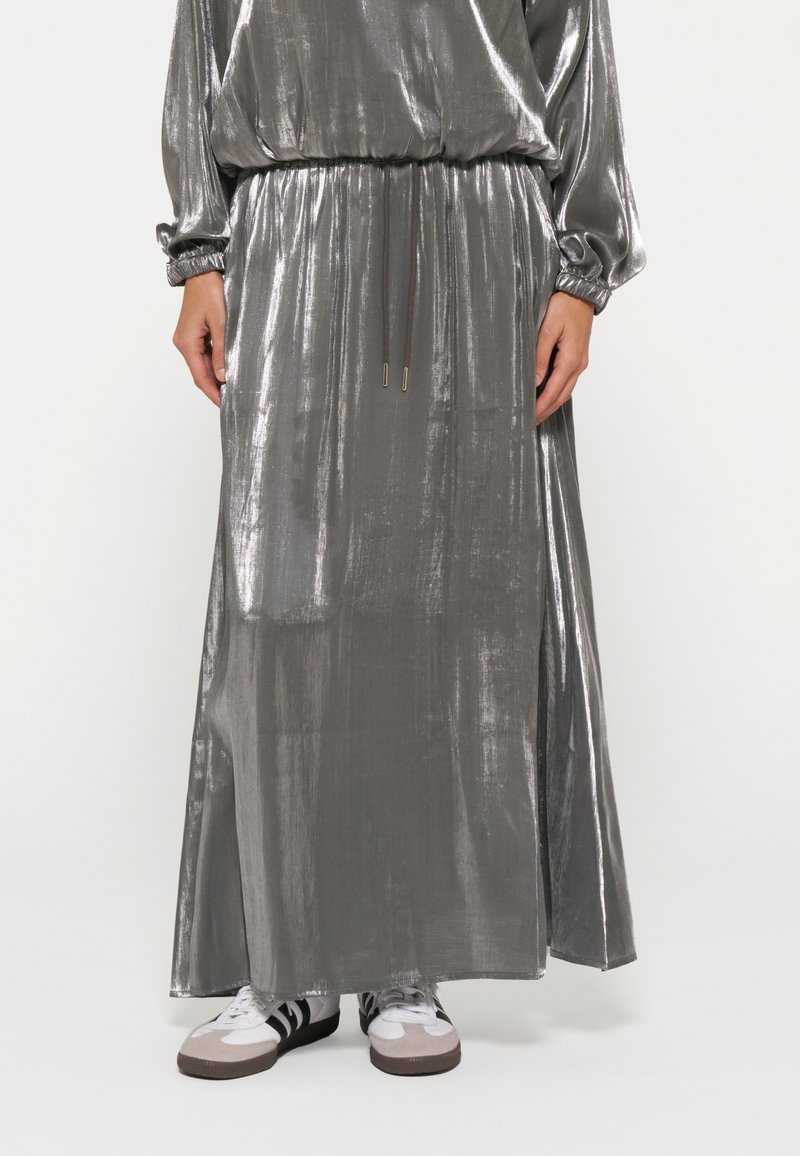 Maxi dress grigio metallizzato con maniche lunghe, vita arricciata e dettaglio con cordoncino. Accostato a calzature bianche e accenti grigi.