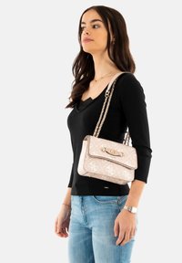 Borsa a mano rosa con un design a quadri, accessori in oro e una tracolla a catena. Indossata a tracolla da una persona che indossa una maglietta nera.