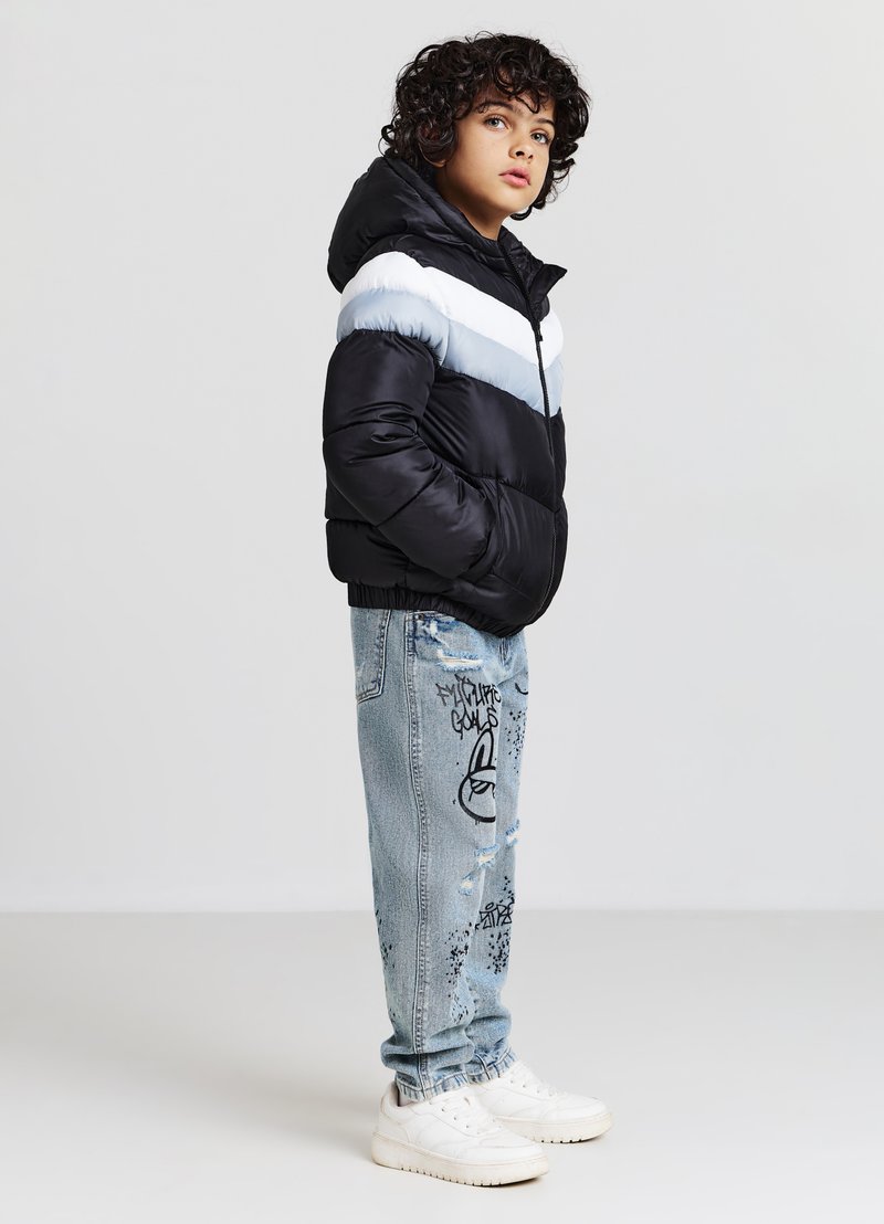 Veste noire bouffante avec des rayures horizontales bleu clair et blanches, associée à un jean bleu clair déchiré orné d'imprimés graphiques noirs et des baskets blanches.