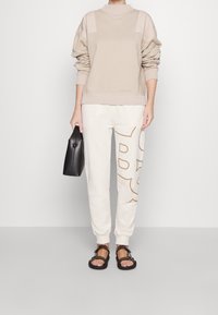 Beiger Pullover mit hellbeigen Schulterpartien, kombiniert mit cremefarbenen Jogginghosen mit braunen Grafikbuchstaben. Schwarze minimalistische Handtasche und Sandalen.