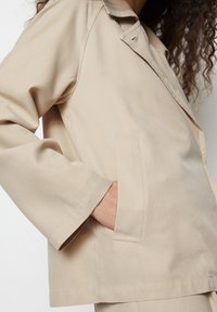 Beżowa kurtka wykonana z miękkiego materiału, z kołnierzem, długimi rękawami, pojedynczą kieszenią na piersi oraz luźnym krojem z przeszyciami.