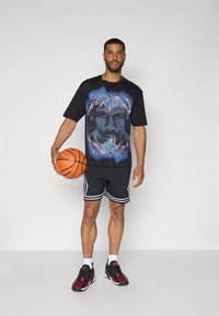 Camiseta gráfica negra con una impresión de cara, combinada con pantalones cortos deportivos negros con franjas laterales blancas y zapatillas rojas y negras. Sosteniendo un balón de baloncesto.