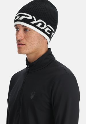 Black knit beanie with white logo band. Textured ribbed design; worn with a black zip-up jacket featuring a subtle spider logo.