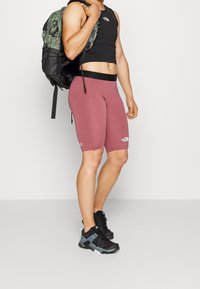 Czarny dopasowany tank top, burgundowe spodenki rowerowe z logo i wielokolorowe obuwie. Zielony plecak w kamuflażu z czarnymi akcentami.