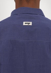 Sötétkék kockás ing gomboló nyakkal, rácsos mintával és hátul egy kontrasztos fekete felirattal ellátott varrott címkével.