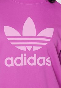 Lila sweatshirt med stor ljusrosa Adidas Trefoil-logotyp och varumärkesnamn på bröstet.