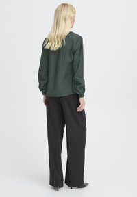 Femeie blondă îmbrăcată cu o bluză lungă cu mâneci verzi închise și pantaloni negri evazați, stând cu spatele pe un fundal deschis.