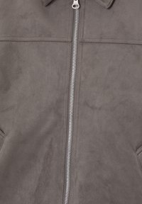 Veste en daim gris avec fermeture éclair centrale en métal et coutures horizontales visibles sur la poitrine et près des poches de chaque côté.
