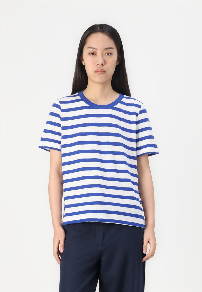 Frau mit langen dunklen Haaren, die ein blau-weiß gestreiftes Kurzarmshirt und dunkle Hosen trägt, steht vor einem schlichten weißen Hintergrund.