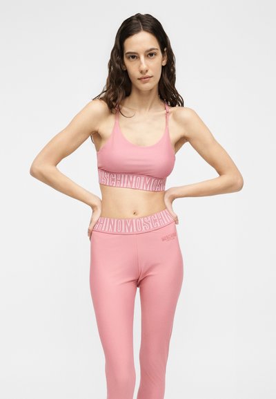 Γυναίκα με κυματιστά καστανά μαλλιά, φορώντας ροζ αθλητικό σουτιέν και κολάν με λευκό λογότυπο Moschino, στέκεται με τα χέρια στη μέση μπροστά σε απλό φόντο.