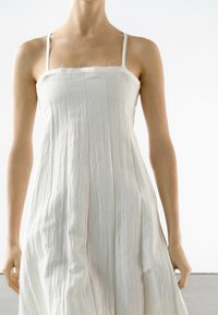 Robe blanche sans manches avec fines bretelles, présentant un design texturé et fluide avec de légers plis. L'ourlet tombe juste au-dessus des genoux.