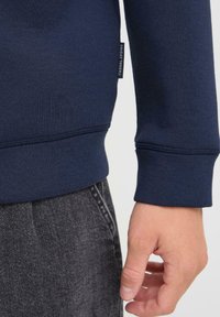 Navy blue sweatshirt fabric with a smooth texture and ribbed cuffs, paired with dark gray denim jeans. Tag visible on the sleeve.
