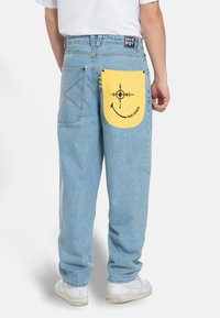 Light blue denim pants with a yellow patch pocket featuring a black graphic design. White sneakers complete the outfit.