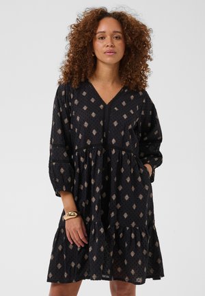 Femme aux cheveux bouclés portant une robe noire à motifs, manches longues et jupe à volants, debout avec une main dans la poche et un bracelet en or au poignet.