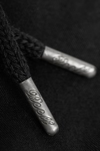 Cadenas de zapatos tejidas en negro con aglets metálicos que presentan un grabado detalle, sobre un fondo oscuro que resalta la textura y la artesanía.