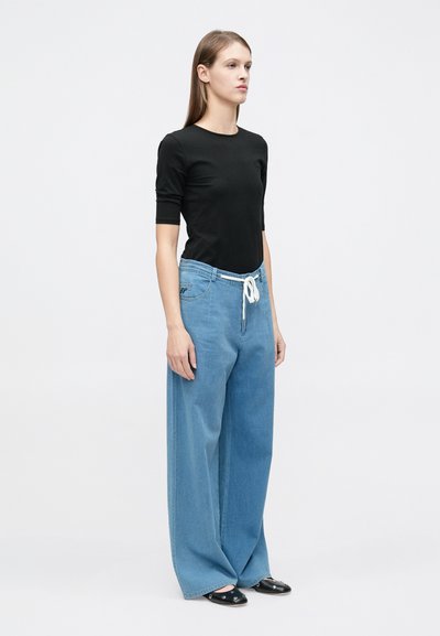 Νεαρή γυναίκα στέκεται, φοράει μαύρο τοπ με μισά μανίκια, ανοιχτό μπλε παντελόνι τζιν με φαρδιά μπατζάκια και λευκό κορδόνι, και μαύρα παπούτσια τύπου slip-on.