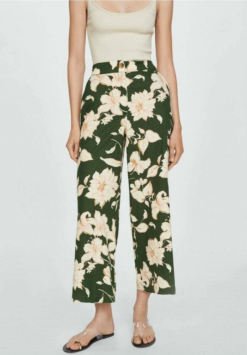 Femme portant un pantalon taille haute à jambes larges vert foncé avec un grand imprimé floral beige, un débardeur beige clair et des sandales à brides transparentes.
