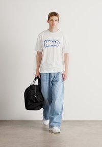 Hvid kortærmet t-shirt med blå "HUGO" tekst, parret med lyseblå vide jeans og hvide sneakers. Bærer en sort duffel bag.