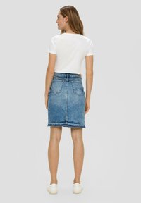Jeansrok met een rafelige zoom, blauwe kleur, twee achterzakken, gedragen met een witte cropped t-shirt en witte sneakers. Effen achtergrond.