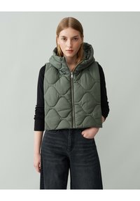 Grüne Steppweste mit Kapuze, Rückenzipper und strukturiertem Muster; getragen über einem schwarzen Langarmshirt und kombiniert mit dunklen Jeans.