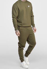 Olivgrön sweatshirt och matchande joggers gjorda av mjukt material. Har rundad halsringning, ribbade ärmmuddar och en liten logotyp på bröstet.